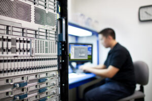 1489031323491 - Assistência Informática - Assistência Informática IT technician sits at a workstation beside a tall server rack, monitoring a monitor in a data center environment.