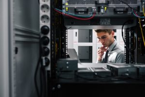 2200-AdobeStock_183267112-1-1024x682 - Assistência Informática - Assistência Informática IT professional inspecting a server rack, looking at a laptop in a data center.