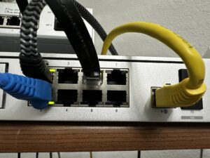 416040575_7823246964358201_2931089499329777331_n - Assistência Informática - Assistência Informática Network switch with blue and yellow Ethernet cables plugged into ports, showing port numbers and cables in use.