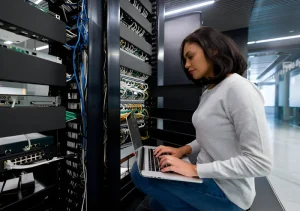 AWS-Cloud-Services-Bahrain - Assistência Informática - Assistência Informática Woman seated by server racks in a data center, typing on a laptop while network cables and equipment loom around her.
