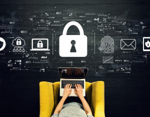 PHYSICAL-IT-SECURITY-SYDNEY - Assistência Informática Person typing on a laptop on a yellow chair, with large padlock and digital security icons floating above, illustrating cybersecurity.