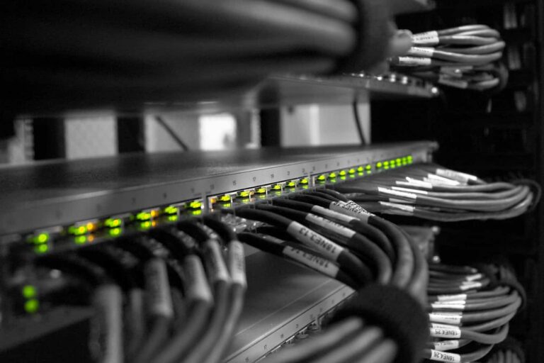 dataroad_rede_informatica - Assistência Informática Network rack with a row of illuminated green LEDs on a switch and bundled Ethernet cables labeled and organized in a server cabinet