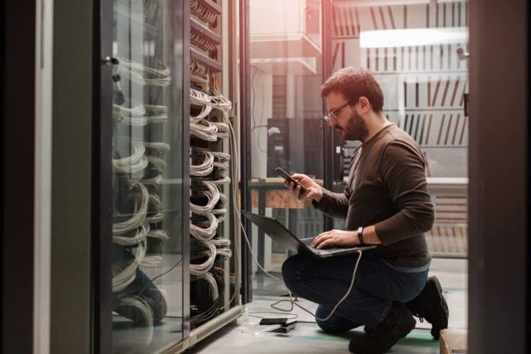 Técnico arrodillado junto a los bastidores de servidores en un centro de datos, usando una computadora portátil y un teléfono inteligente.