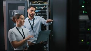 images3 - Assistência Informática Two IT professionals in a data center review a laptop beside server racks, with one gesturing toward the equipment.