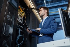 it-support-what-is-it-and-do-you-need-it-750x500 - Assistência Informática - Assistência Informática IT professional in a blue suit inspecting server racks with a laptop in a data center