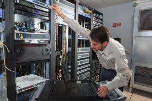 man_datacenter - Assistência Informática - Assistência Informática Technician in a data center reaching to rack servers, with a laptop on a rolling cart nearby and network cables in the racks.