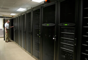 technician_working - Assistência Informática - Assistência Informática IT technician in a white shirt stands by a row of tall black server racks in a data center corridor, door open to a cabinet.