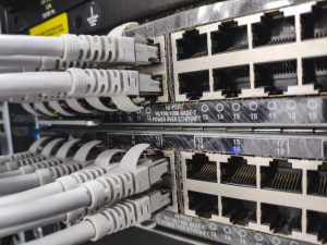 panel switch with free ports - Assistência Informática - Assistência Informática Close-up of a network patch panel with multiple RJ-45 Ethernet ports and gray cables plugged into them in a server rack, showing data cabling.