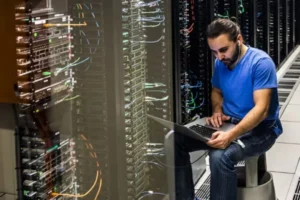 wireless - Assistência Informática - Assistência Informática Tech worker in a blue shirt sits beside server racks, using a laptop in a data center to troubleshoot.