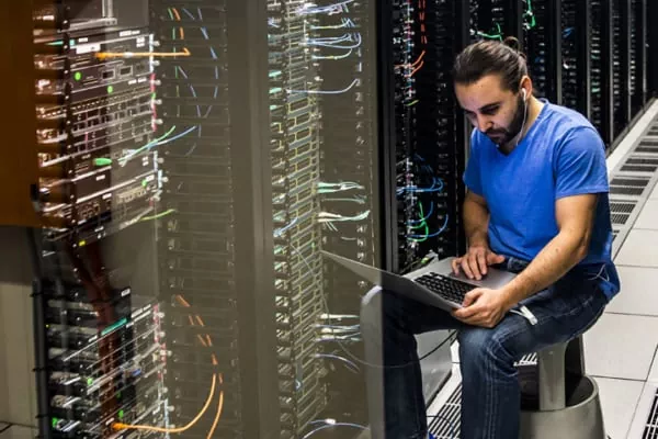 Trabajador tecnológico con camisa azul se sienta junto a bastidores de servidores, usando una computadora portátil en un centro de datos para solucionar problemas.